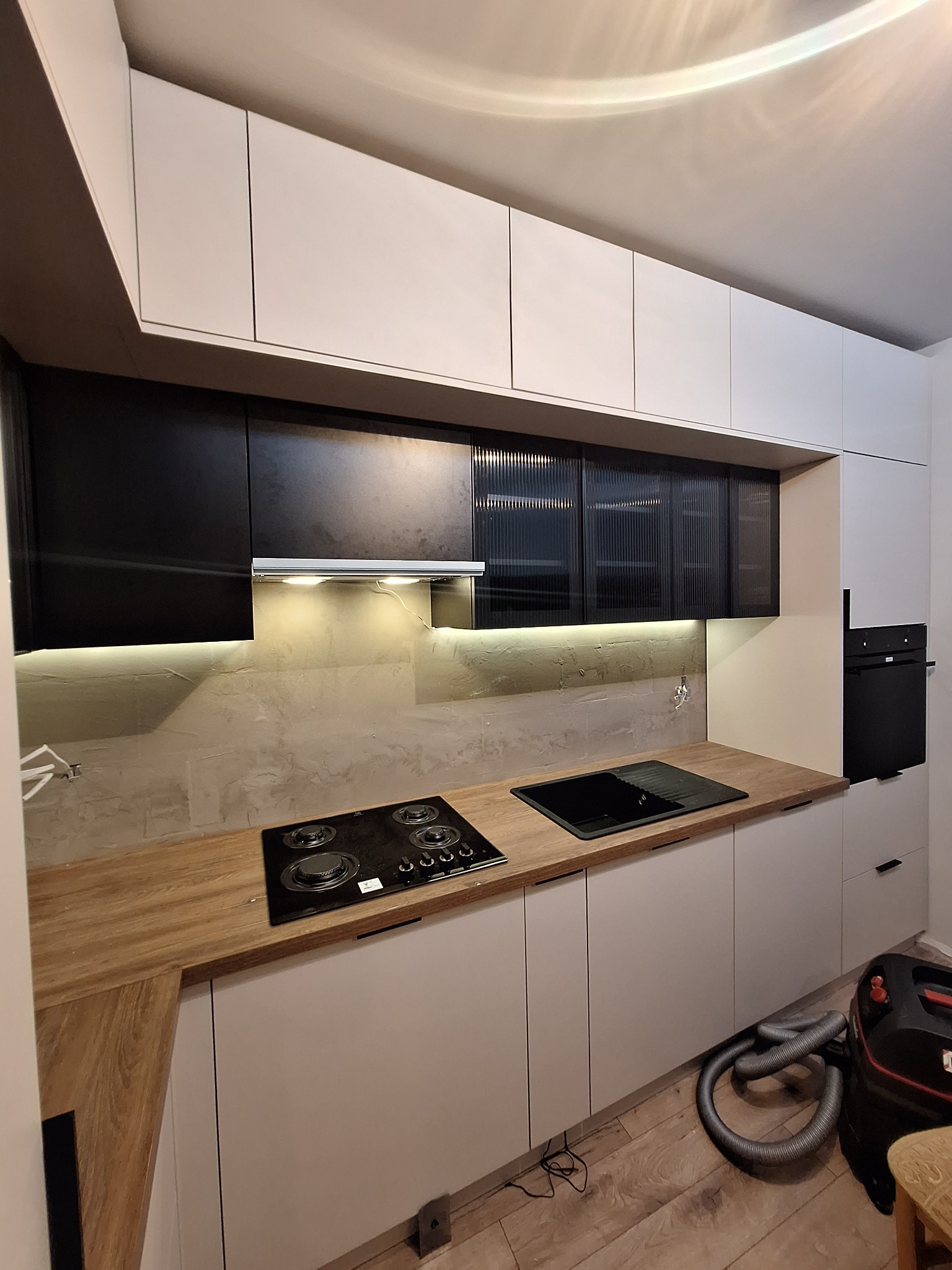 Nowoczesna kuchnia z białymi szafkami dolnymi i czarnymi górnymi, drewniany blat, płyta grzewcza i zlew. Widoczny odkurzacz. Oświetlenie LED pod szafkami.