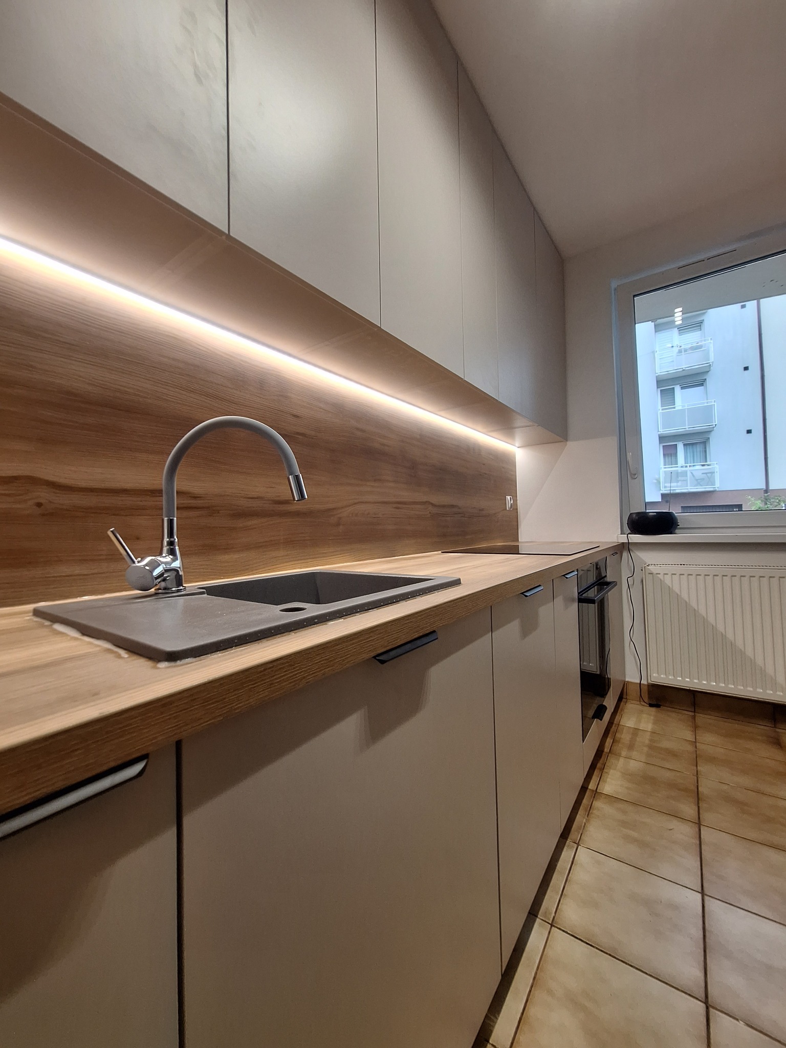 Nowoczesna kuchnia z beżowymi szafkami, drewnianym blatem i podświetleniem LED. Widok na okno z widokiem na budynek mieszkalny. Minimalistyczny design.