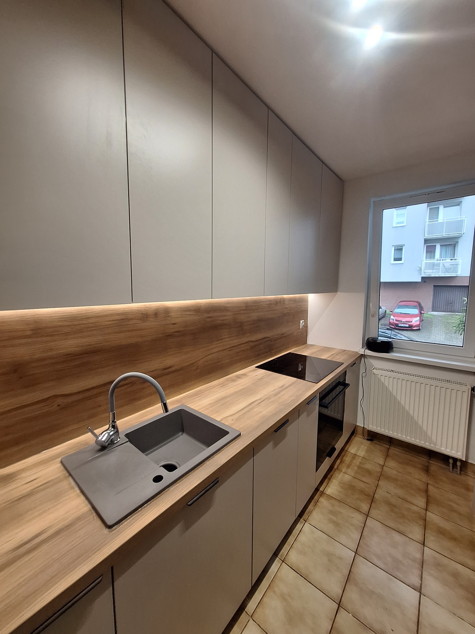 Nowoczesna kuchnia z szarymi frontami, drewnianym blatem i podświetleniem LED. Widok na okno z widokiem na budynek i czerwony samochód. Minimalistyczny design.