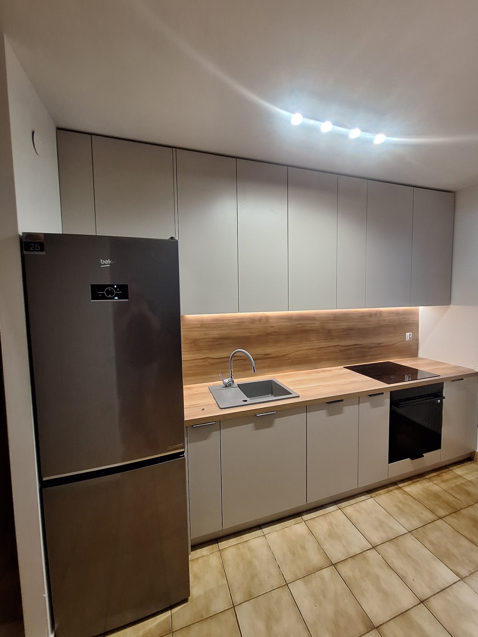 Nowoczesna kuchnia z szarymi szafkami, drewnianym blatem i srebrną lodówką. Oświetlenie LED pod szafkami górnymi. Płytki podłogowe w odcieniach beżu i brązu.