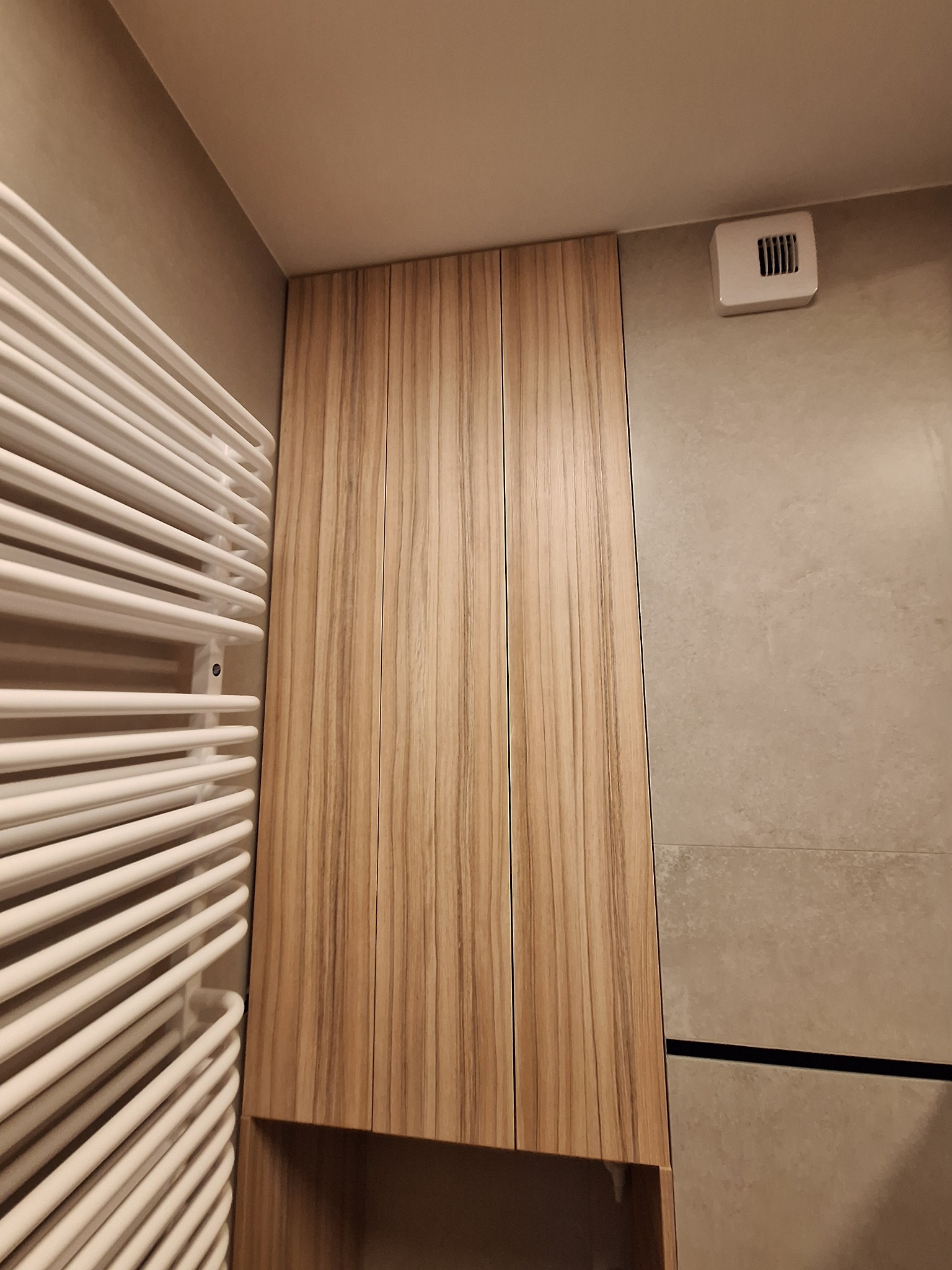 Drewniana szafka łazienkowa na wymiar z widocznym grzejnikiem drabinkowym i wentylacją. Ujęcie z dołu, podkreślające wysokość zabudowy.