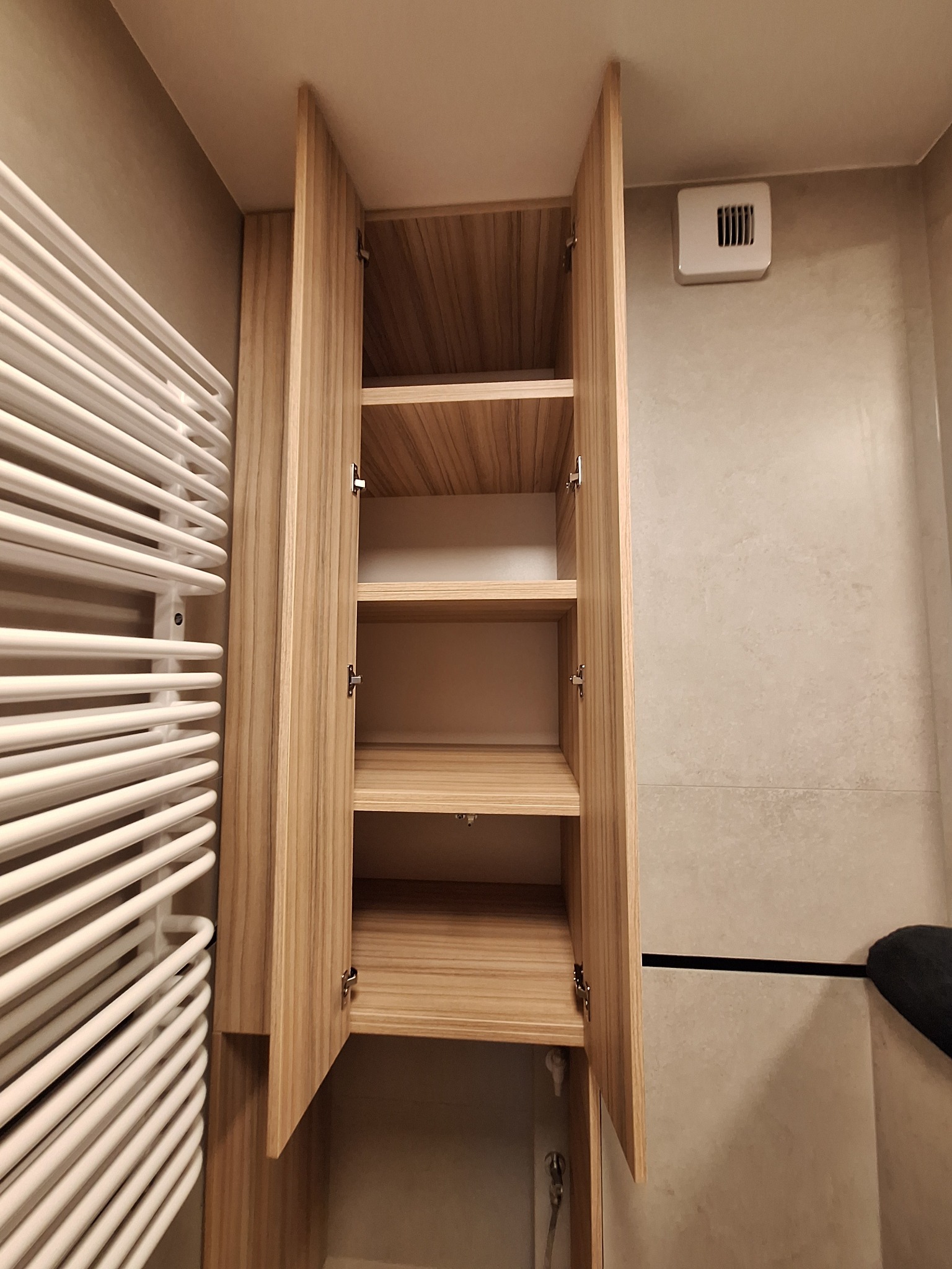 Otwarta, jasna szafka łazienkowa z drewnianymi półkami, zamontowana obok białego grzejnika drabinkowego na beżowej ścianie z wentylacją.