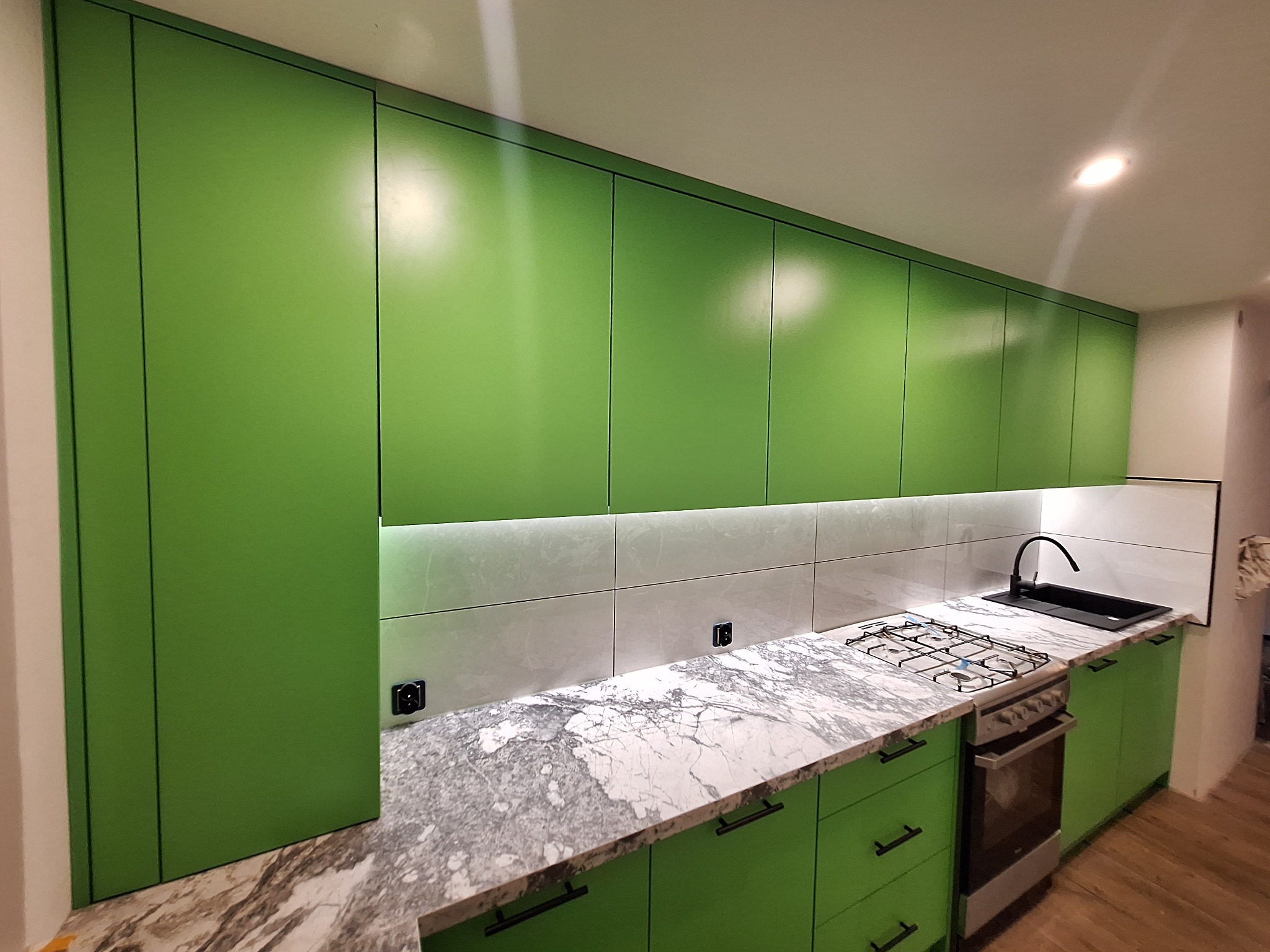 Nowoczesna kuchnia na wymiar z zielonymi frontami, marmurowym blatem i białymi płytkami. Górne szafki podświetlone LED. Widoczna płyta gazowa i czarny zlew.