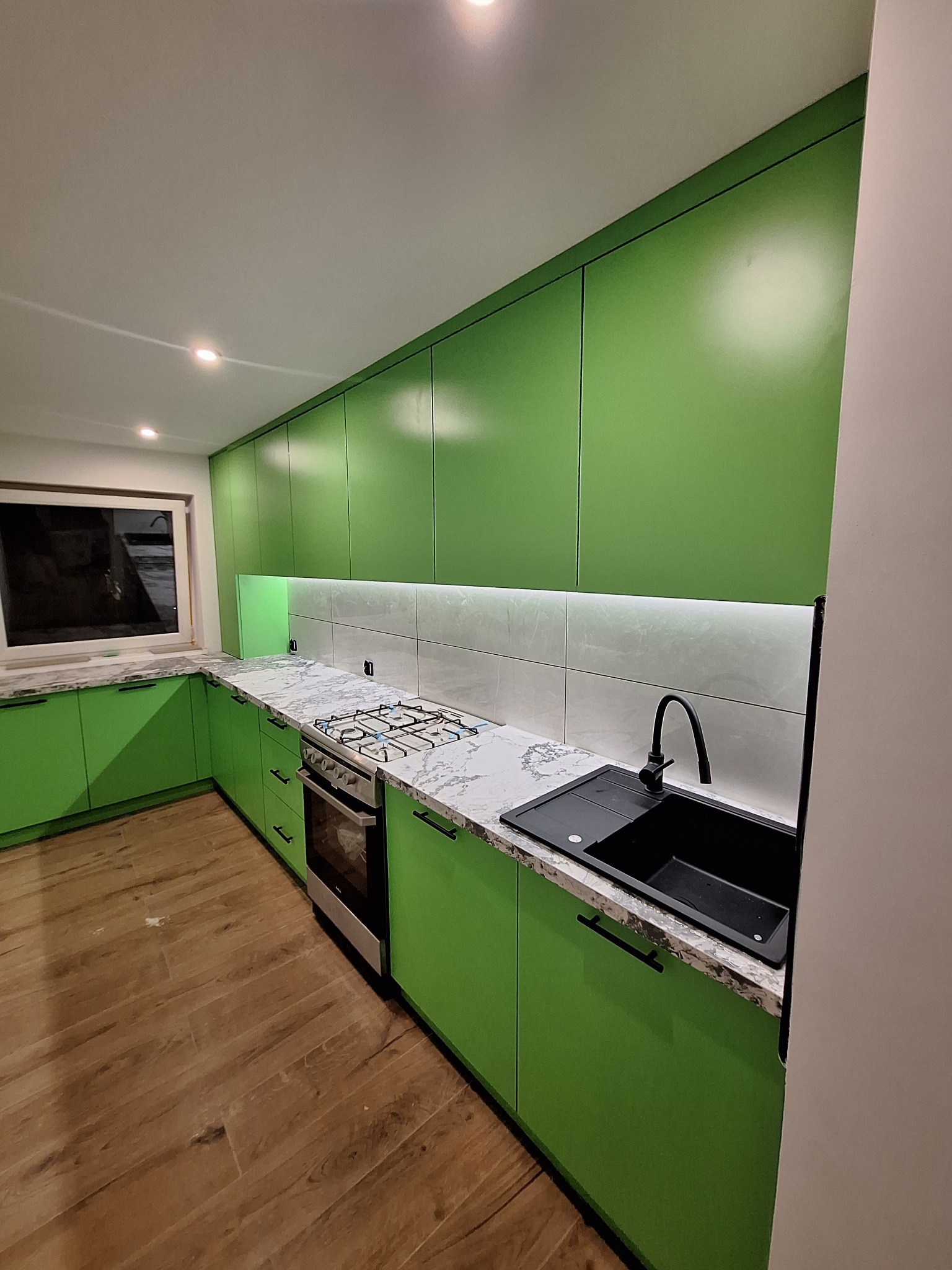Nowoczesna kuchnia z zielonymi frontami, marmurowym blatem i czarnym zlewem. Uwagę przykuwa oświetlenie LED pod szafkami wiszącymi i drewniana podłoga.