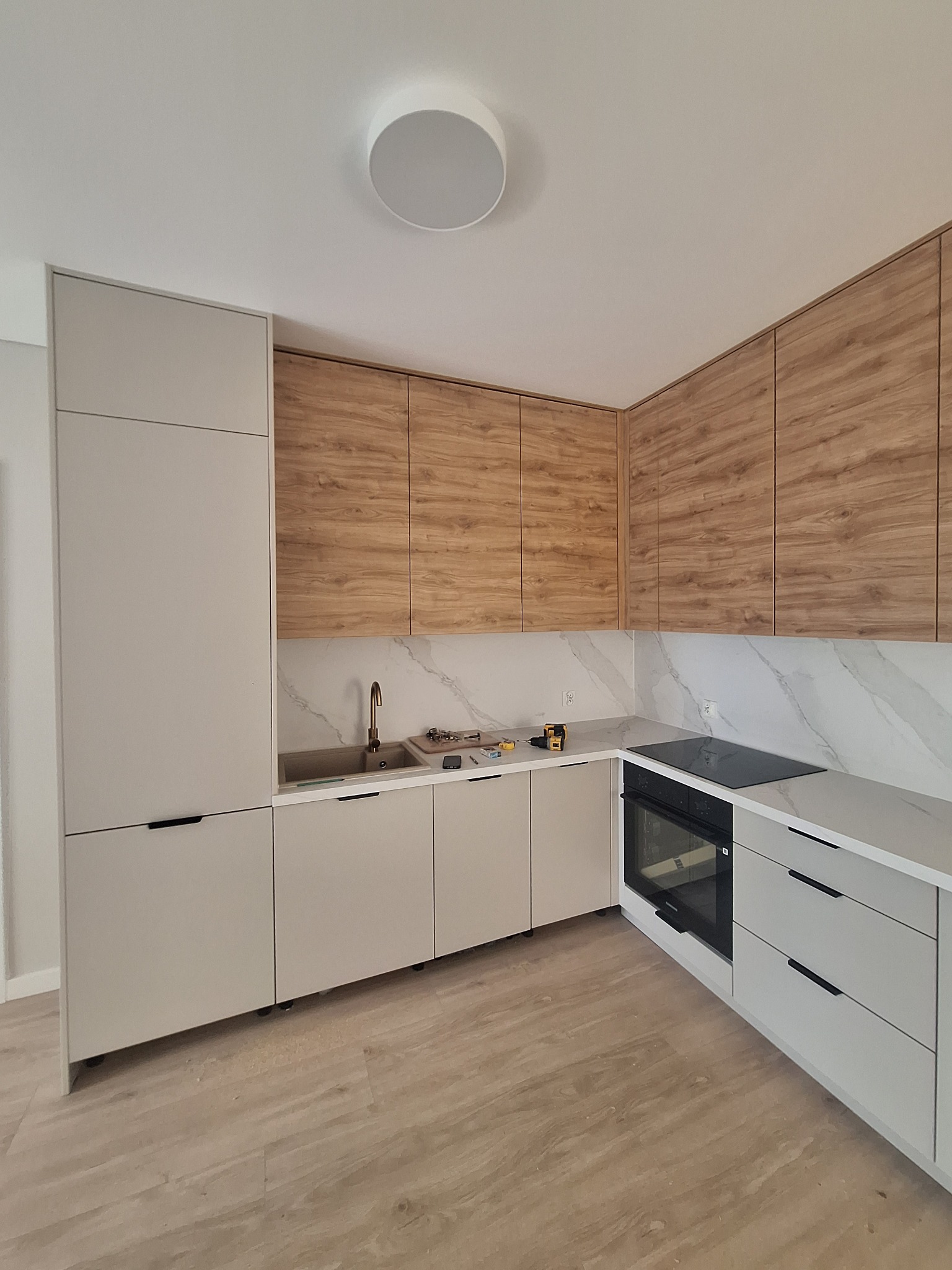 Jasna kuchnia z drewnianymi szafkami wiszącymi i marmurowym blatem. Na blacie złota bateria, płyta indukcyjna i narzędzia monterskie. Nowoczesny design.