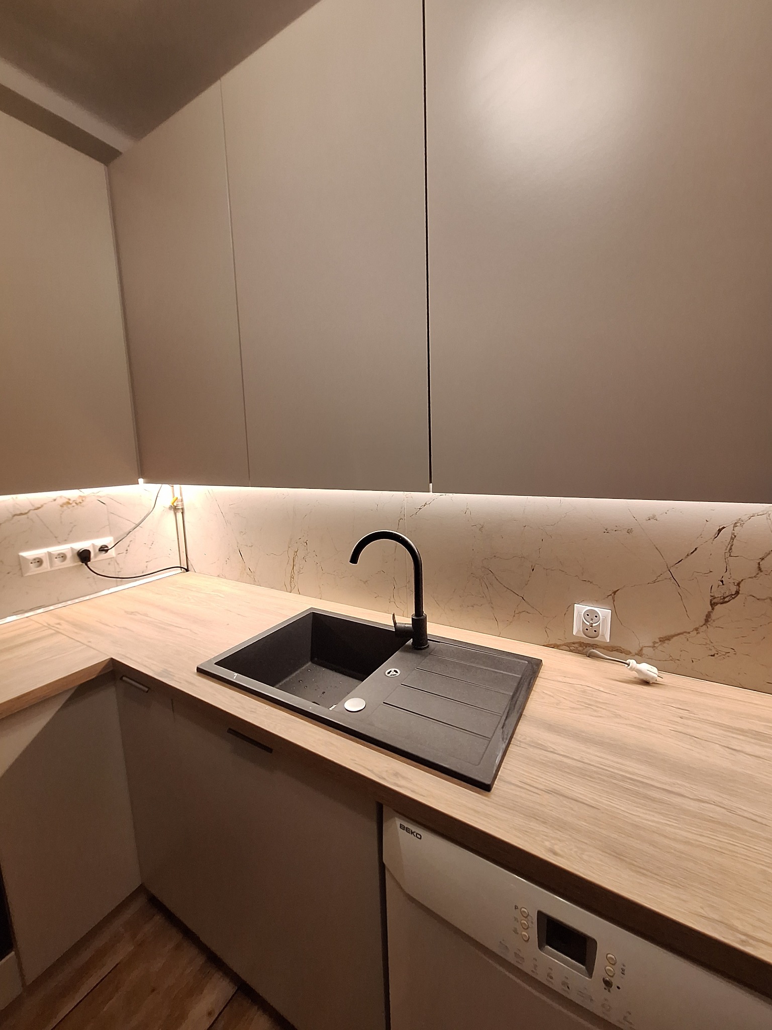 Nowoczesna kuchnia z beżowymi frontami, drewnianym blatem i czarnym zlewem. Podświetlenie LED nad blatem. Widoczna zmywarka Beko. Minimalistyczny design.