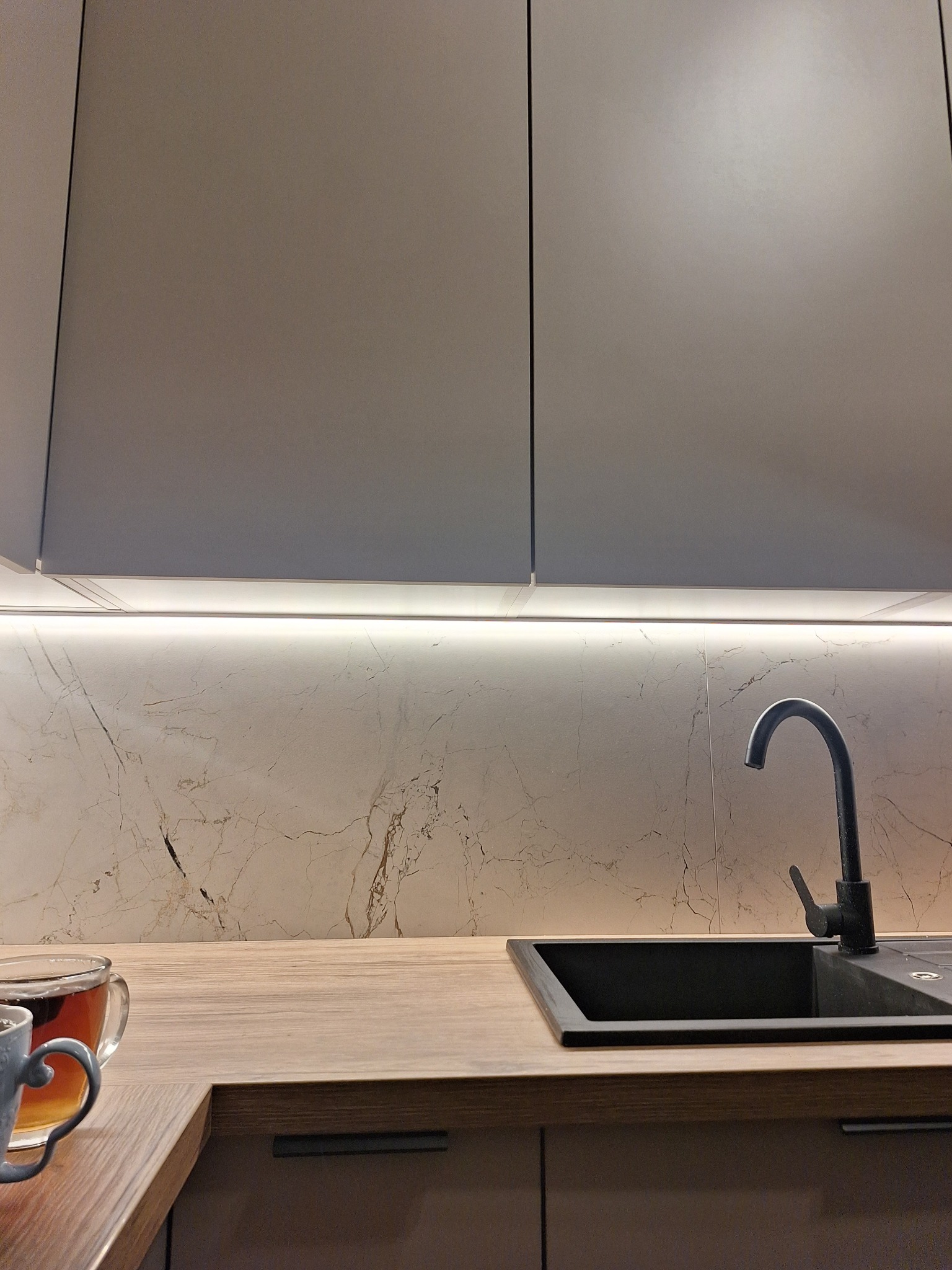 Nowoczesna kuchnia: szare szafki, marmurowy backsplash z oświetleniem LED, czarny zlew i bateria. Na blacie dwie filiżanki z herbatą.