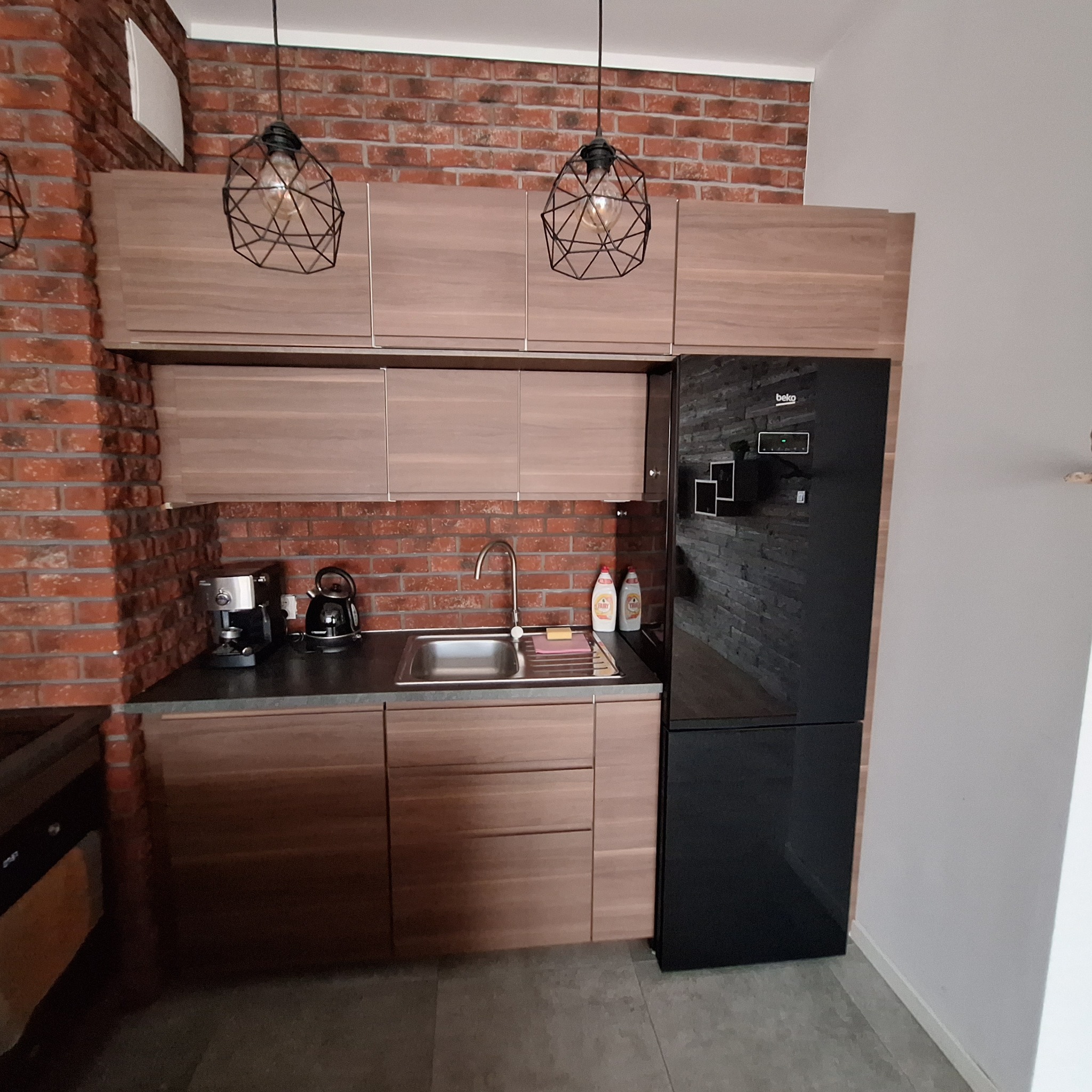 Nowoczesna zabudowa kuchenna z drewnianymi frontami i ceglaną ścianą w tle. Minimalistyczne lampy wiszące. Realizacja z Katowic.