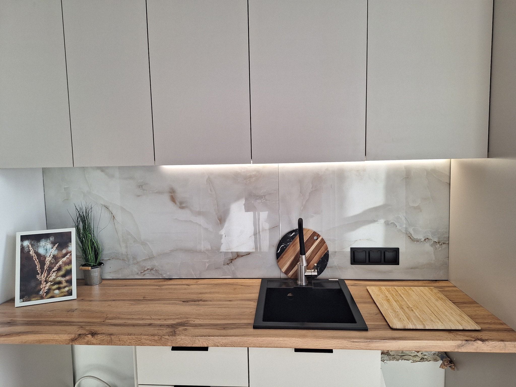 Nowoczesna kuchnia z białymi szafkami, drewnianym blatem i marmurowym panelem. Uwagę przykuwa czarny zlew i okrągła deska do krojenia z elementami drewna.