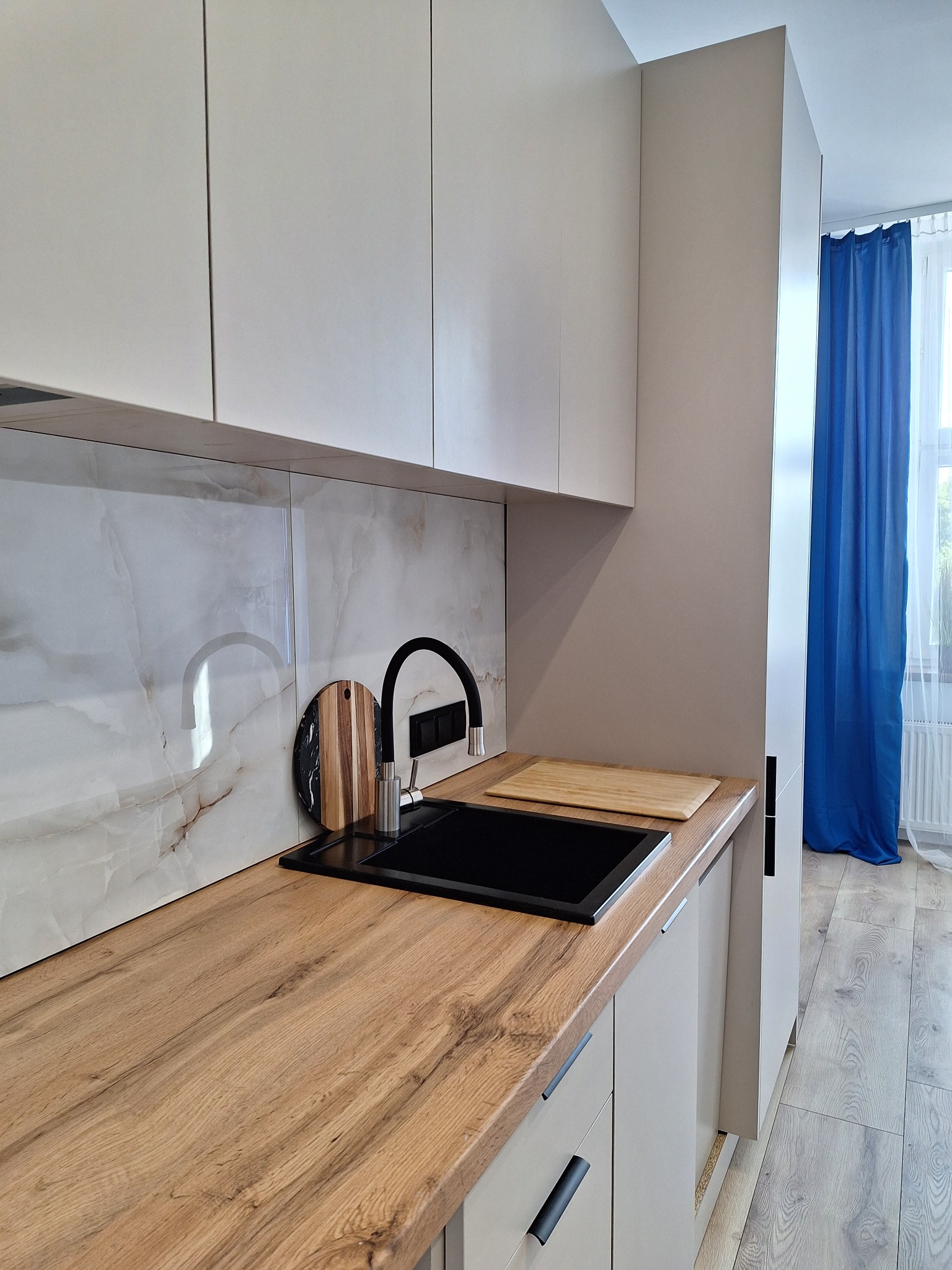 Jasna kuchnia z drewnianym blatem, czarnym zlewem i marmurową ścianą. Uwagę przyciąga niebieska zasłona w oknie, dodająca koloru minimalistycznemu wnętrzu.