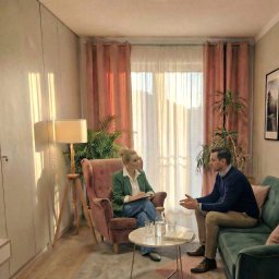 INTROS SPÓŁKA Z OGRANICZONĄ ODPOWIEDZIALNOŚCIĄ - Sesja terapeutyczna w jasnym gabinecie: kobieta w zielonej marynarce notuje, mężczyzna gestykuluje siedząc na zielonej sofie. Na stoliku woda i książka.