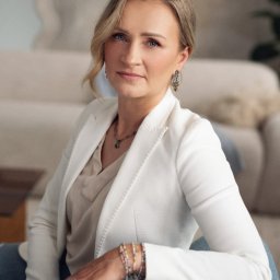 INTROS SPÓŁKA Z OGRANICZONĄ ODPOWIEDZIALNOŚCIĄ - Portret kobiety w białej marynarce, siedzącej na fotelu w jasnym wnętrzu. Uwagę przyciągają delikatne kolczyki i bransoletki na jej ręce.