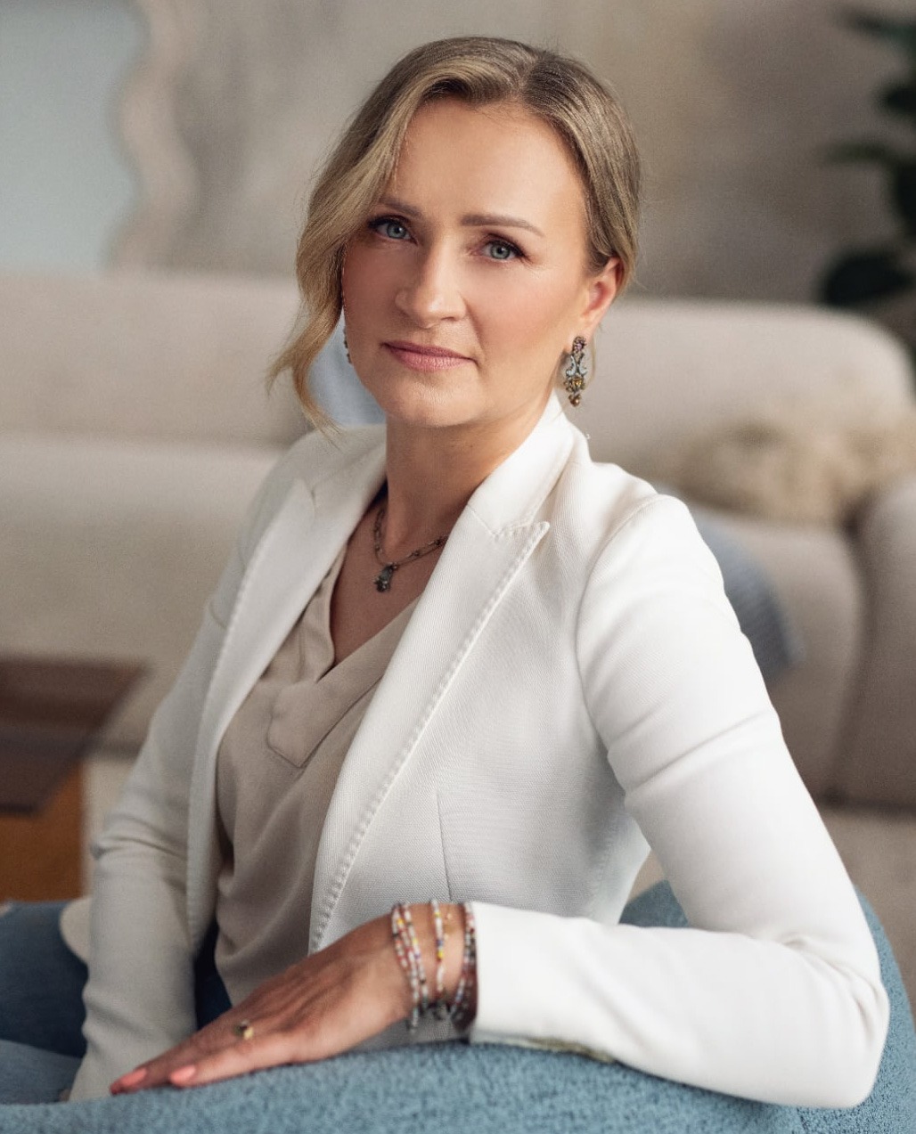 Portret kobiety w białej marynarce, siedzącej na fotelu w jasnym wnętrzu. Uwagę przyciągają delikatne kolczyki i bransoletki na jej ręce.