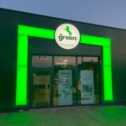 Fasada budynku z logo 'Green Unicorn Group' podświetlonym na zielono. W tle widoczne plakaty reklamowe pomp ciepła i klimatyzacji.