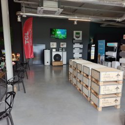 Wnętrze salonu z ekspozycją klimatyzatorów różnych marek, pomp ciepła i akcesoriów. Na pierwszym planie palety z zapakowanymi urządzeniami.