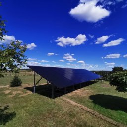 Instalacja fotowoltaiczna na trawniku w słoneczny dzień. Panele ustawione pod kątem, widoczne drzewa i błękitne niebo z chmurami. Ujęcie z dołu.