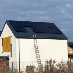 GREEN UNICORN GROUP SPÓŁKA Z OGRANICZONĄ ODPOWIEDZIALNOŚCIĄ - Biały dom z panelami słonecznymi na dachu i drabiną opartą o ścianę. Widok z dołu, w tle pochmurne niebo i fragment sąsiedniego budynku.