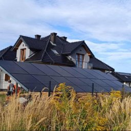 GREEN UNICORN GROUP SPÓŁKA Z OGRANICZONĄ ODPOWIEDZIALNOŚCIĄ - Instalacja fotowoltaiczna na gruncie przed domem jednorodzinnym z czarną dachówką, widoczna ekipa montażowa i trawa na pierwszym planie. Błękitne niebo w tle.