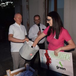 GREEN UNICORN GROUP SPÓŁKA Z OGRANICZONĄ ODPOWIEDZIALNOŚCIĄ - Losowanie nagród: kobieta w różowej koszulce wyciąga los z ozdobnego kosza, obok mężczyźni z mikrofonem i koszem, w tle voucher z logo Green Unicorn Group.