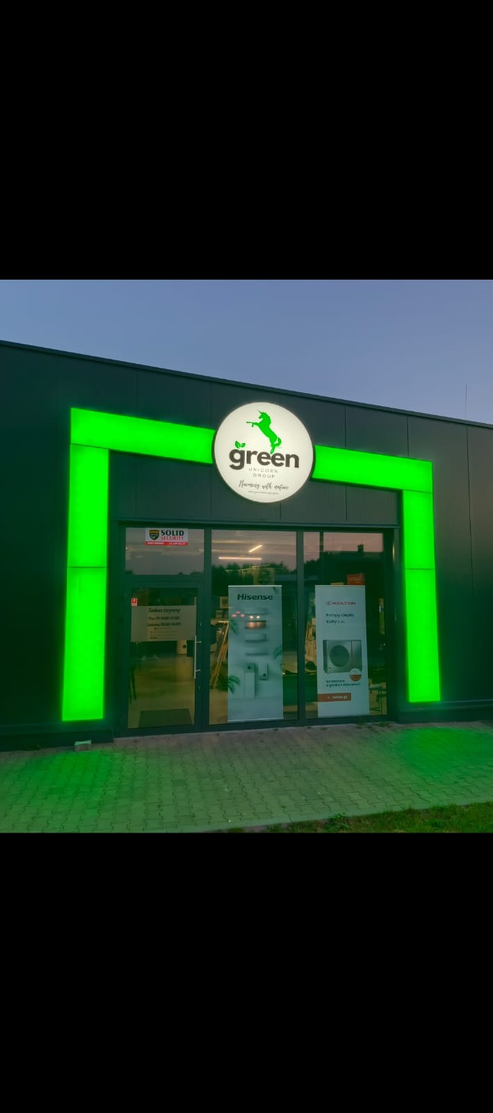 Fasada budynku z logo 'Green Unicorn Group' podświetlonym na zielono. W tle widoczne plakaty reklamowe pomp ciepła i klimatyzacji.