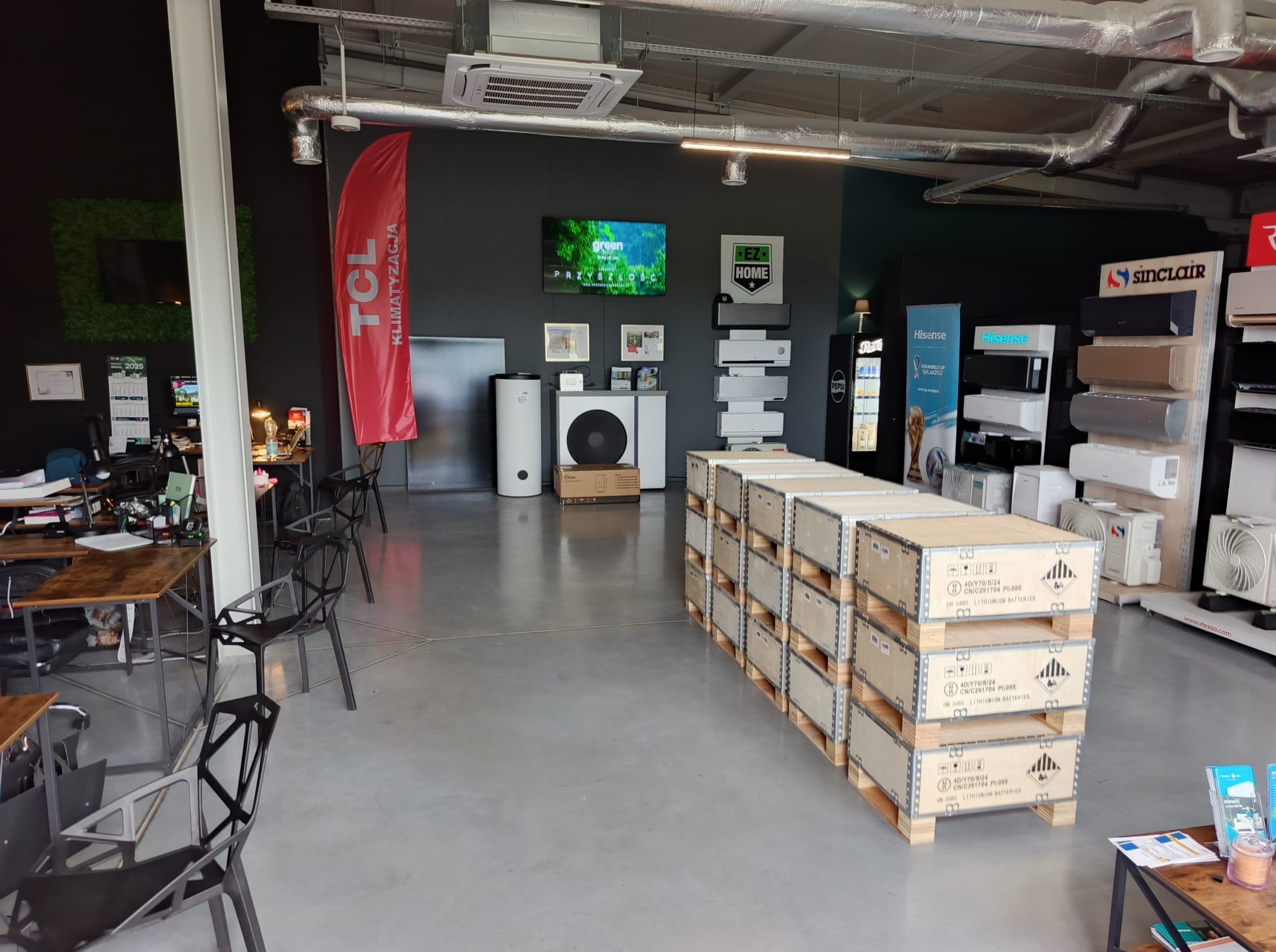 Wnętrze salonu z ekspozycją klimatyzatorów różnych marek, pomp ciepła i akcesoriów. Na pierwszym planie palety z zapakowanymi urządzeniami.