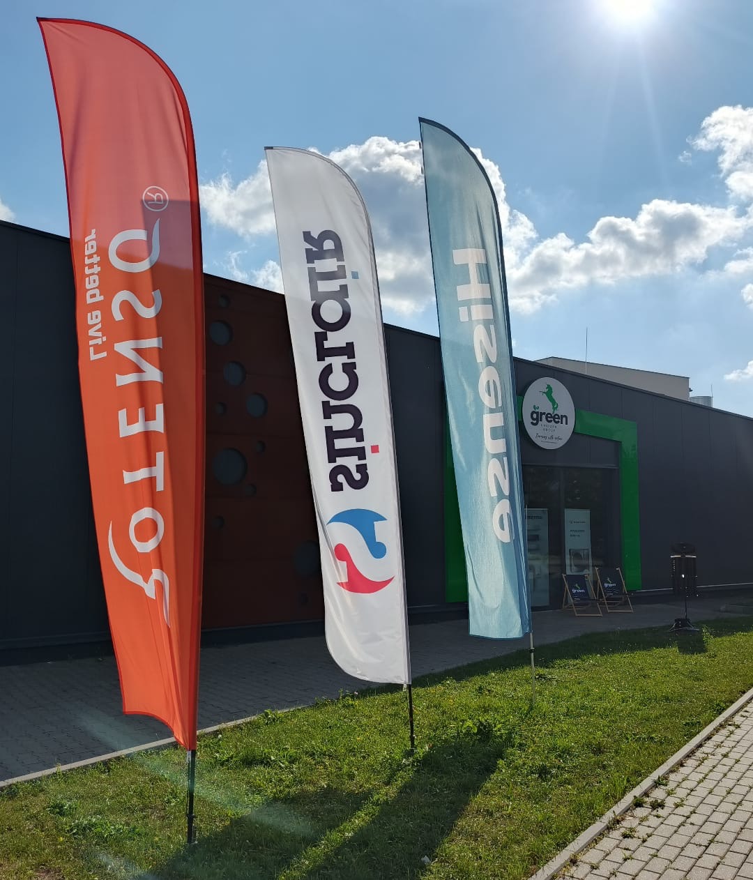 Trzy flagi reklamowe przed budynkiem z logo 'green' na tle błękitnego nieba z chmurami. Flagi w kolorach: pomarańczowym, białym i błękitnym.