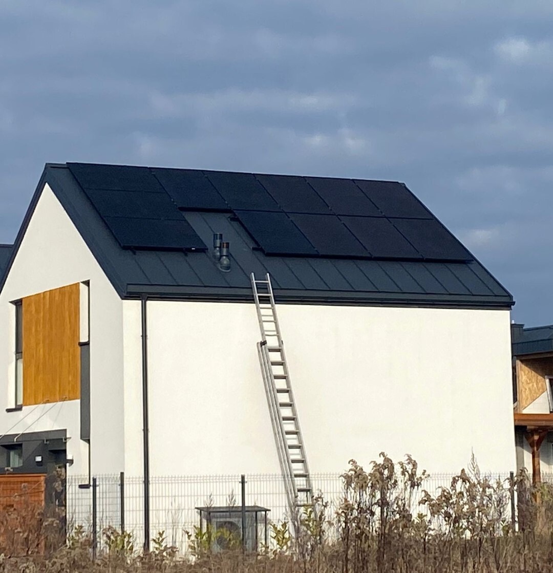 Biały dom z panelami słonecznymi na dachu i drabiną opartą o ścianę. Widok z dołu, w tle pochmurne niebo i fragment sąsiedniego budynku.