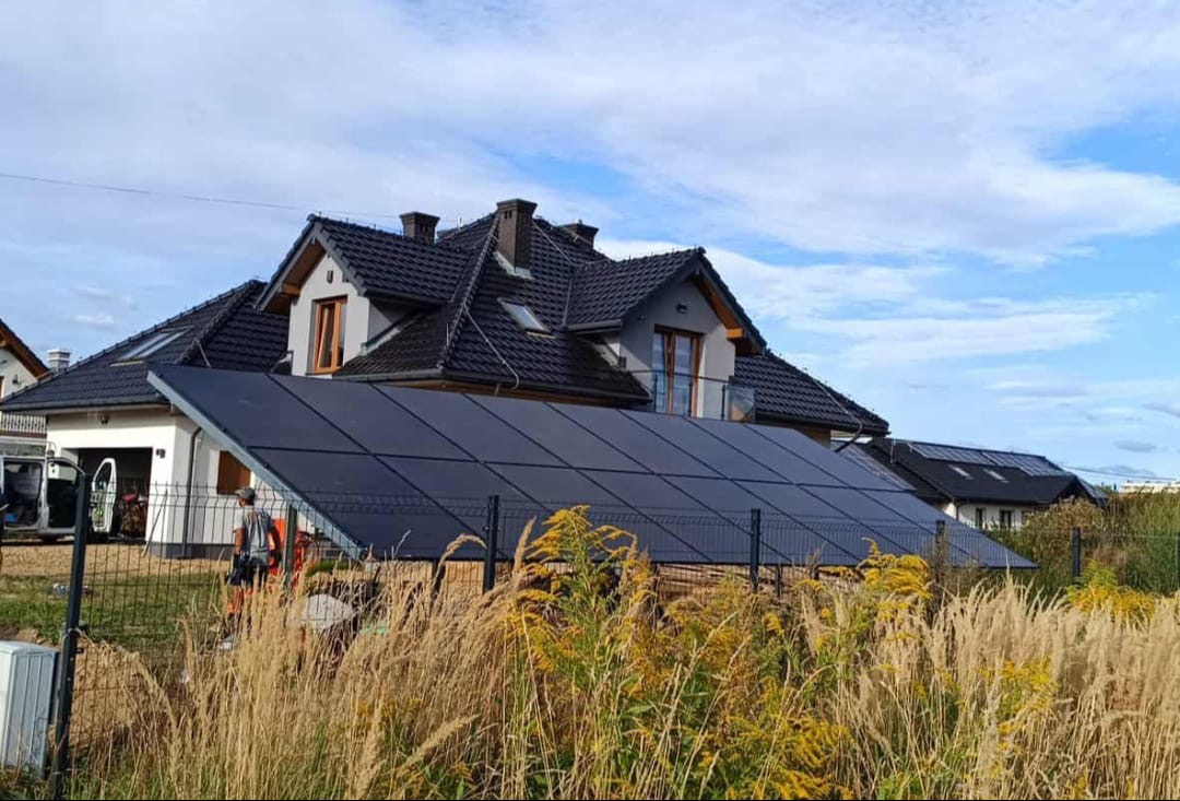 Instalacja fotowoltaiczna na gruncie przed domem jednorodzinnym z czarną dachówką, widoczna ekipa montażowa i trawa na pierwszym planie. Błękitne niebo w tle.