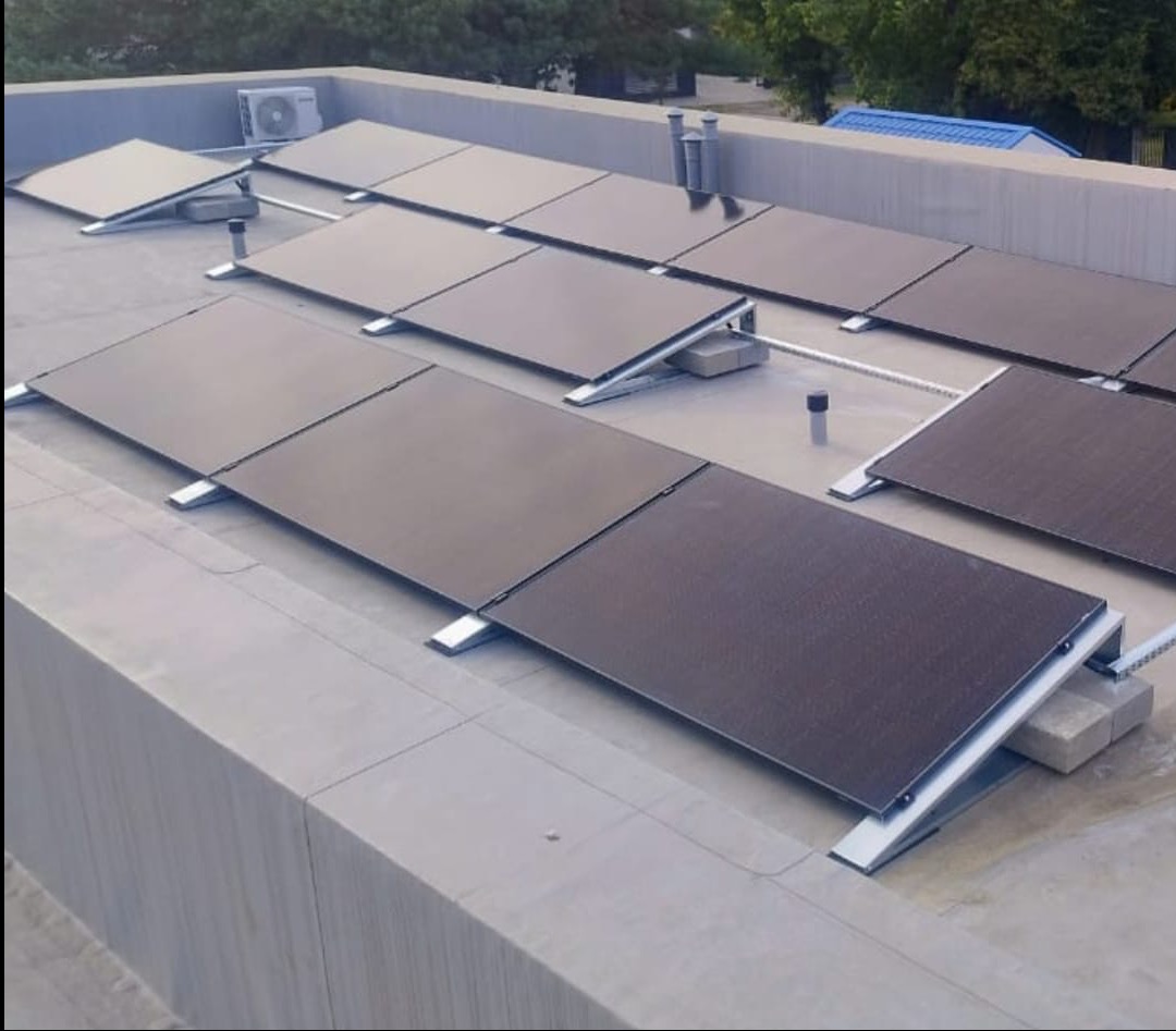 Instalacja fotowoltaiczna na płaskim dachu budynku z widoczną klimatyzacją. Panele zamontowane na aluminiowych konstrukcjach, betonowe podpory. Widok z góry, lekko z boku.