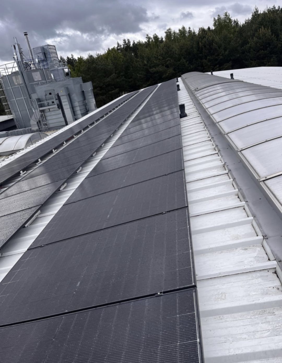 Rząd paneli fotowoltaicznych na białym, metalowym dachu budynku przemysłowego, z widocznym systemem wentylacyjnym i lasem w tle, pochmurne niebo.
