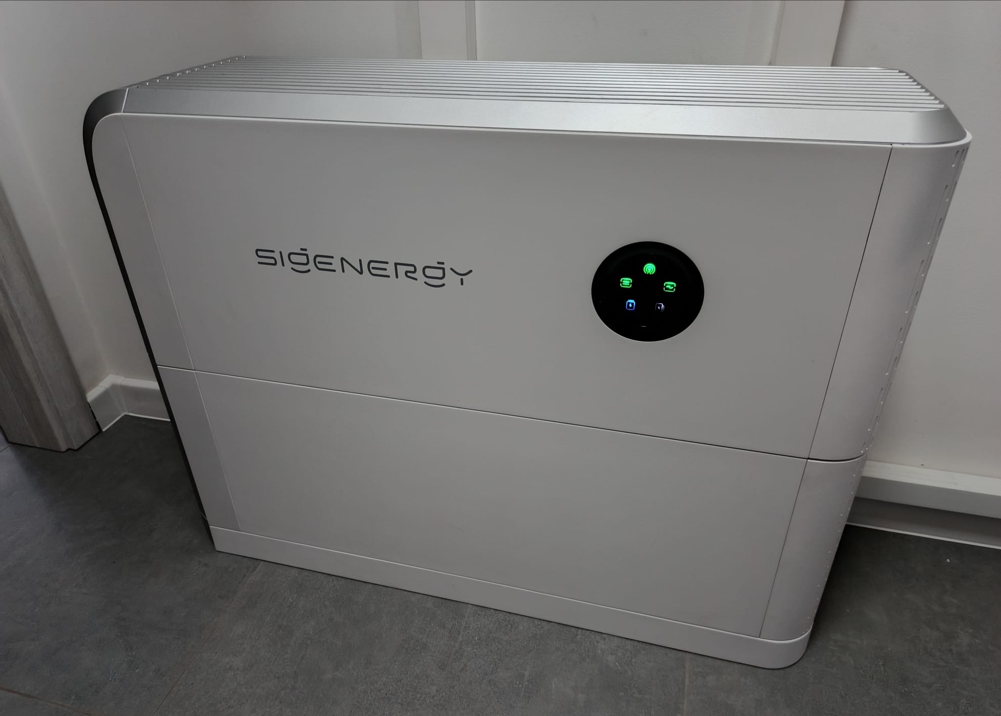 Biały, nowoczesny akumulator energii SiGenergy z okrągłym panelem sterowania i zielonymi diodami, ustawiony na szarej podłodze w jasnym pomieszczeniu.