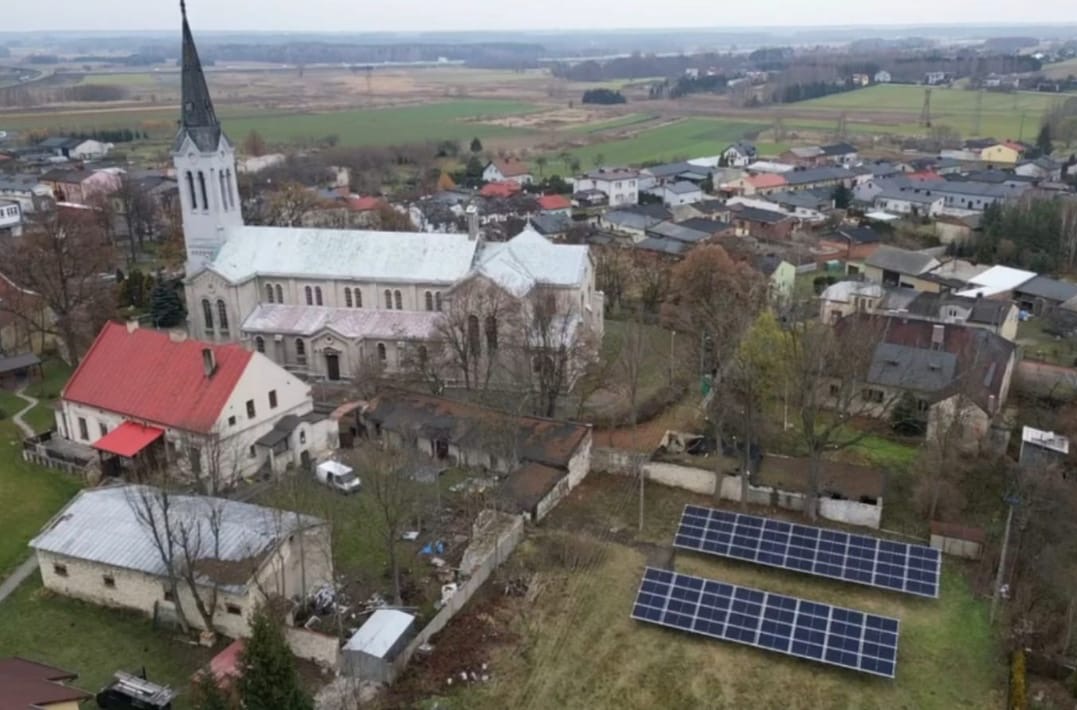 Widok z góry na panele fotowoltaiczne w Katowicach, zainstalowane obok zabudowań z kościołem w tle. Ujęcie z drona ukazuje całą instalację w krajobrazie.