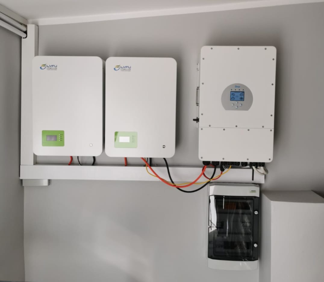 Białe akumulatory energii i inwerter fotowoltaiczny LVFU zamontowane na ścianie, z widoczną skrzynką bezpiecznikową. Instalacja wewnątrz pomieszczenia.