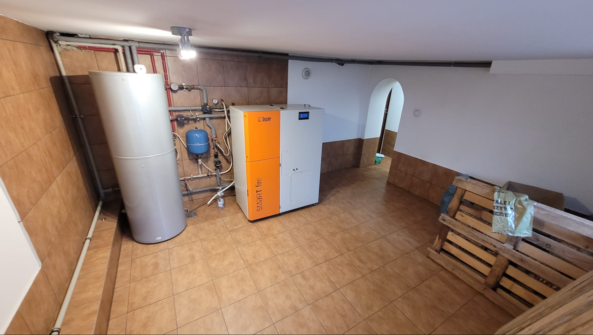 Kotłownia z piecem Lazar Smart Fire, zbiornikiem i instalacją. Pomieszczenie wyłożone płytkami, widoczne rury i osprzęt. Ujęcie z perspektywy podłogi.