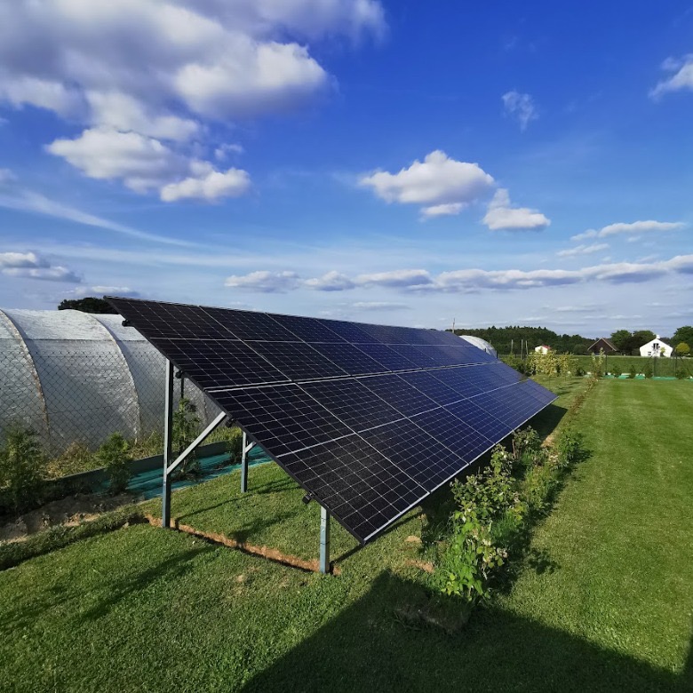 Panel fotowoltaiczny na trawniku, z widoczną szklarnią w tle, pod błękitnym niebem z białymi obłokami. Instalacja na metalowej konstrukcji, otoczona zielenią.