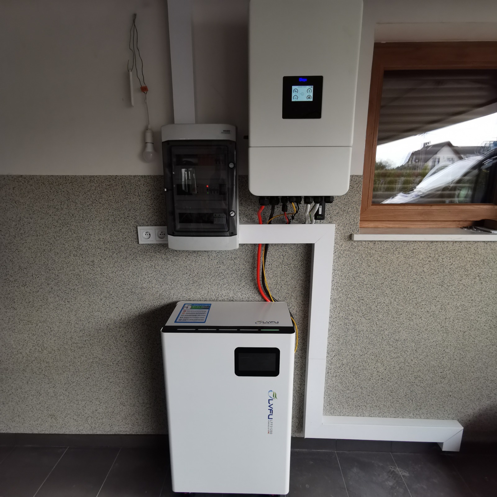 Biały akumulator energii i falownik fotowoltaiczny zamontowane na ścianie w pomieszczeniu. Widoczne kable i elementy instalacji elektrycznej.