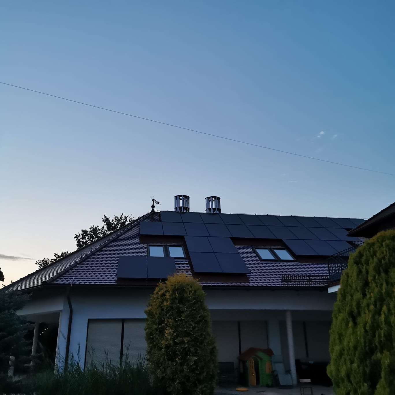 Dom jednorodzinny z panelami fotowoltaicznymi na dachu, widoczne kominy i okna dachowe. Ujęcie z dołu, w tle błękitne niebo o zmierzchu.