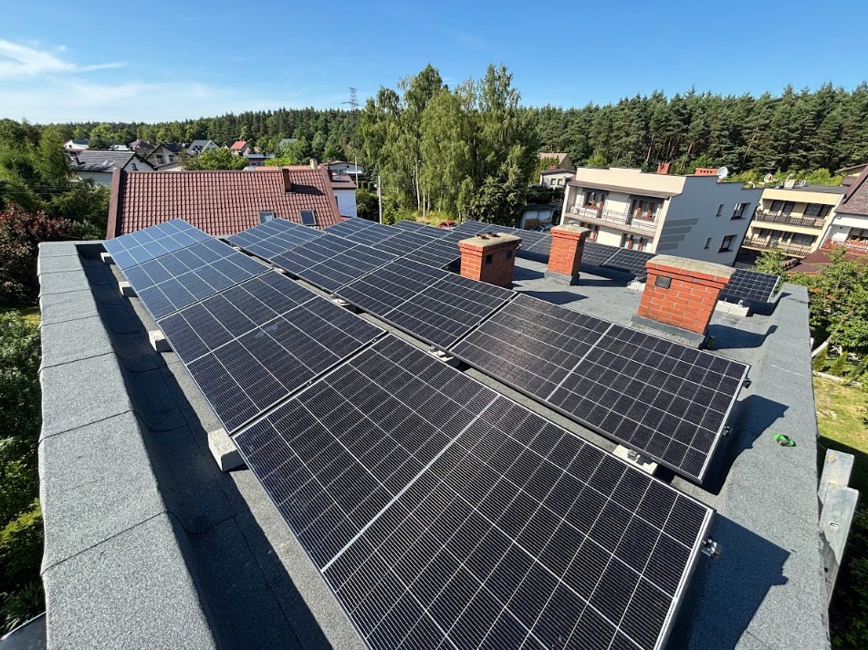 Panele fotowoltaiczne na dachu domu jednorodzinnego, widok z góry, w tle zabudowa mieszkalna i las, słoneczny dzień.