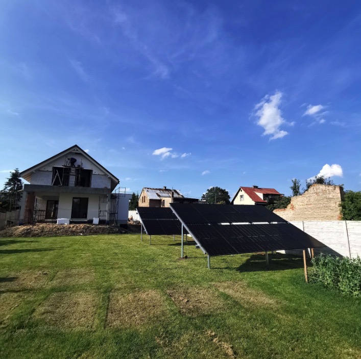 Montaż paneli fotowoltaicznych na trawniku przed domem w trakcie budowy, błękitne niebo. Widoczne rusztowania i prace wykończeniowe na elewacji.