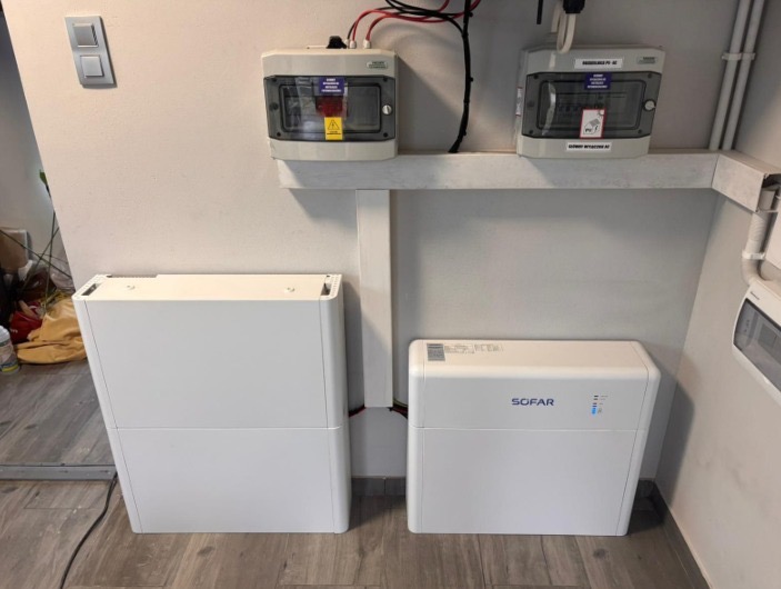 Dwa białe akumulatory Sofar obok siebie, podłączone do instalacji elektrycznej z zabezpieczeniami. Widoczne kable i skrzynki elektryczne na ścianie.