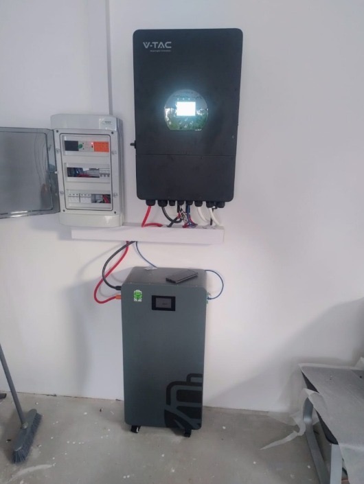 Czarny inwerter V-TAC na ścianie nad szarą baterią akumulatorów z podłączonymi kablami. Obok skrzynka elektryczna. Elementy instalacji fotowoltaicznej w pomieszczeniu.