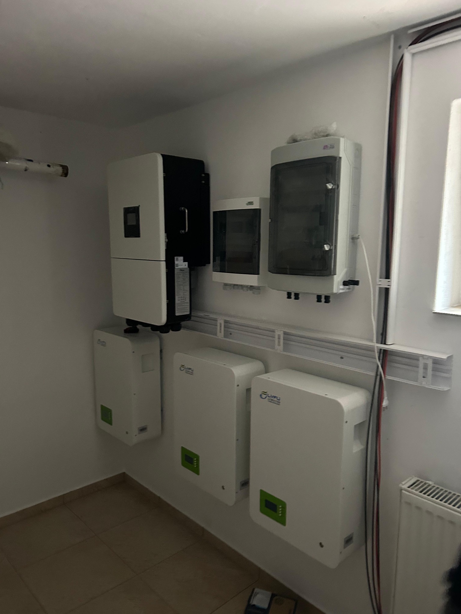 Kompletna instalacja fotowoltaiczna w pomieszczeniu: inwerter, rozdzielnica i akumulatory, zamontowane na ścianie, widoczne okablowanie i grzejnik.