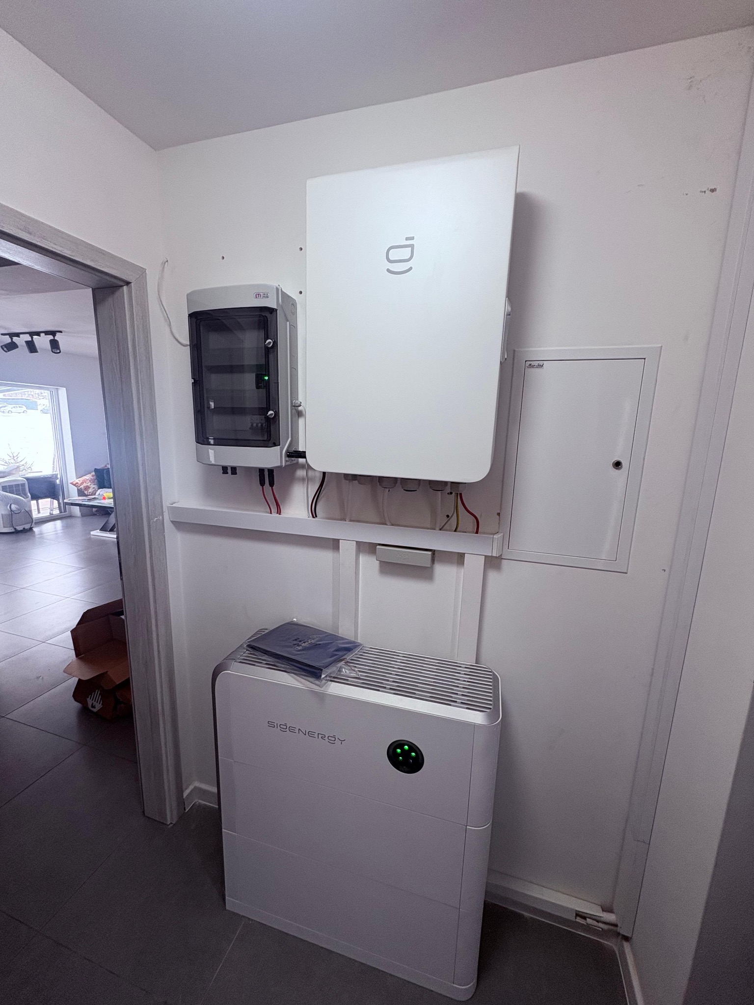 Wnętrze z zamontowaną białą pompą ciepła SIGENERGY, rozdzielnicą elektryczną i skrzynką. Widoczne przewody i elementy instalacji. Minimalistyczny design.