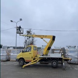 GIFTED Ewa Dmochowska - Żółty samochód z wysięgnikiem i pracownikiem w koszu, naprawiającym oświetlenie uliczne. Widoczne palety z materiałami w tle.