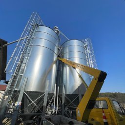 GIFTED Ewa Dmochowska - Dwa srebrne silosy zbożowe z drabinkami, oświetlone słońcem, z żółtym ramieniem podnośnika koszowego z napisem 'Wynajem 507 793 127' na tle błękitnego nieba.