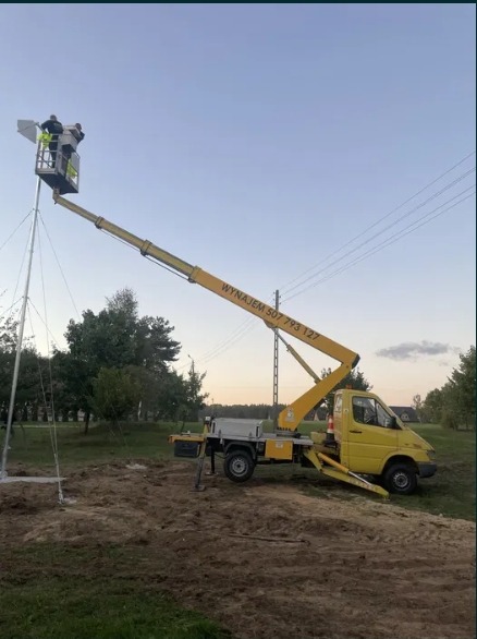 Żółty pojazd z wysięgnikiem i dwoma pracownikami w koszu, montujący konstrukcję na zewnątrz. Widoczne linie energetyczne i drzewa w tle, zmierzch.
