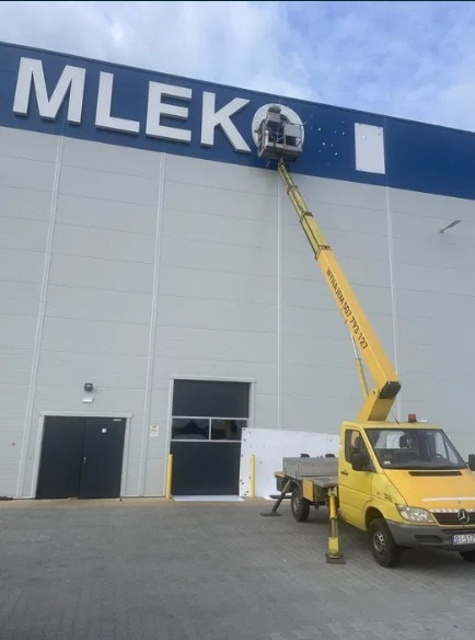 Żółty pojazd z wysięgnikiem przy elewacji budynku z napisem 'MLEKO', osoba w koszu wysięgnika wykonuje prace na wysokości.