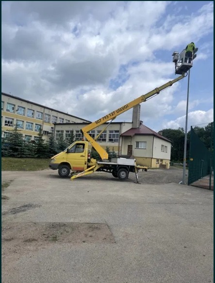 Żółty pojazd z wysięgnikiem i koszem na wysokości oświetla lampę uliczną. W tle budynek z oknami i pochmurne niebo. Numer telefonu na ramieniu wysięgnika.
