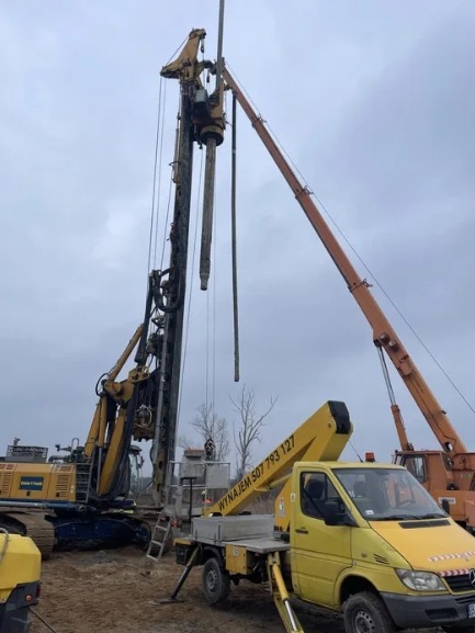 Żółty podnośnik koszowy z wysięgnikiem na tle wiertnicy i pomarańczowego dźwigu. Prace wysokościowe w trakcie realizacji, widoczny numer kontaktowy na pojeździe.