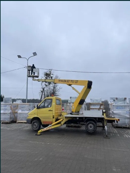 Żółty samochód z wysięgnikiem i pracownikiem w koszu, naprawiającym oświetlenie uliczne. Widoczne palety z materiałami w tle.